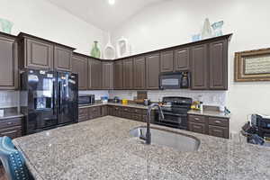 Kitchen featuring dark brown cabinetry, black appliances, decorative backsplash, light stone counters, and vaulted ceiling