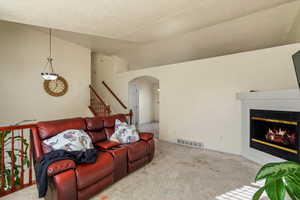 Carpeted living room with a glass covered fireplace, arched walkways, vaulted ceiling, stairway, and a textured ceiling