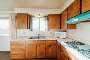 Kitchen featuring brown cabinetry, stainless steel gas cooktop, white microwave, light countertops, and under cabinet range hood