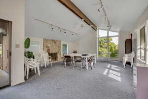 Dining space featuring rail lighting, beamed ceiling, light colored carpet, a stone fireplace, and plenty of natural light