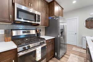 Kitchen with stainless steel appliances, light wood-style floors, decorative backsplash, dark brown cabinets, and light stone counters