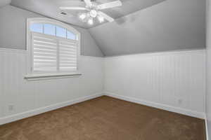 Bonus room with lofted ceiling, ceiling fan, dark colored carpet, and wainscoting