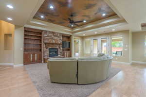 Living room with built in shelves, light wood finished floors, a raised ceiling, ceiling fan, and a stone fireplace