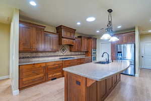 Kitchen featuring stainless steel appliances, light stone counters, pendant lighting, an island with sink, and light wood-style flooring