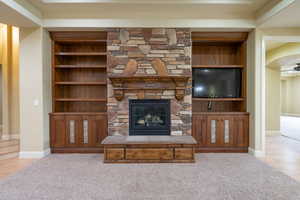 Unfurnished living room with built in features, a stone fireplace, ceiling fan, and light colored carpet