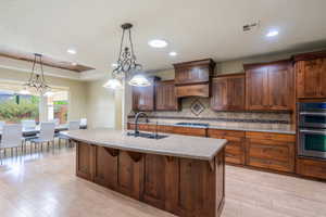 Kitchen with hanging light fixtures, stainless steel appliances, tasteful backsplash, a center island with sink, and light stone countertops