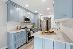 Kitchen with appliances with stainless steel finishes, light stone counters, blue cabinets, light wood-style floors, and decorative backsplash