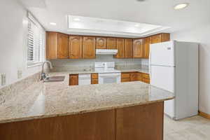 Kitchen featuring brown cabinetry, white appliances, a peninsula, light stone counters, and a tray ceiling