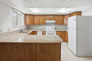 Kitchen featuring white appliances, brown cabinetry, light stone countertops, a peninsula, and a raised ceiling