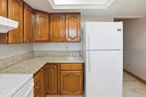 Kitchen featuring white appliances, brown cabinets, light stone counters, and range hood