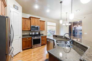 Kitchen featuring appliances with stainless steel finishes, brown cabinetry, hanging light fixtures, light wood-type flooring, and dark stone counters