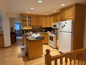 Kitchen with glass insert cabinets, white appliances, a kitchen island, brown cabinets, and dark countertops