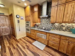 Kitchen featuring brown cabinetry, wall chimney exhaust hood, a warming drawer, dark wood-style flooring, and recessed lighting