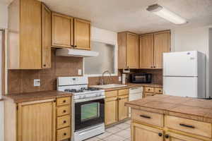 Kitchen featuring white appliances, light tile patterned floors, tile counters, under cabinet range hood, and a textured ceiling