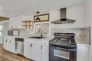 Kitchen with appliances with stainless steel finishes, wall chimney exhaust hood, white cabinetry, backsplash, and open shelves
