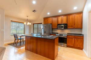 Kitchen featuring wood cabinets, stainless steel appliances, a center island with sink, dark countertops, and hanging light fixtures, at counter sitting.