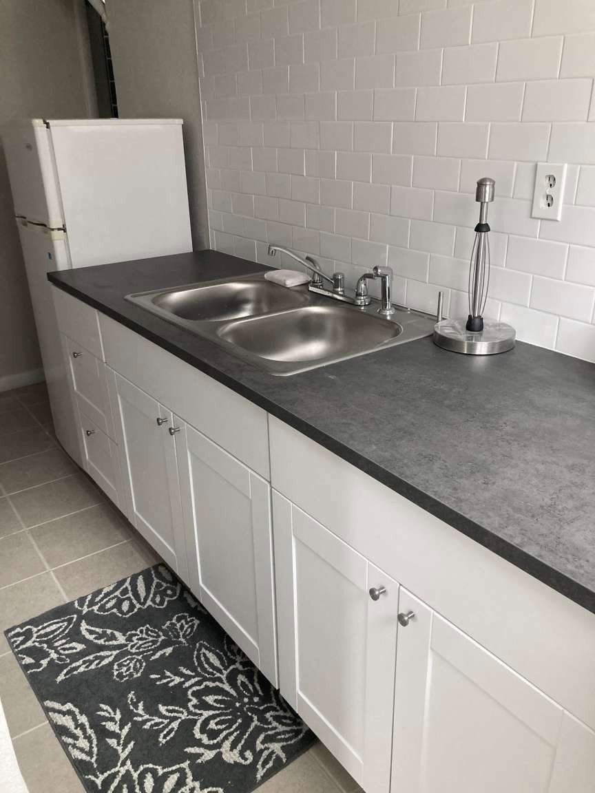 Kitchenette featuring a double basin stainless steel sink, solid surface countertop, white shaker style cabinetry, and subway tile backsplash