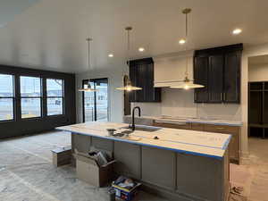 Kitchen featuring a center island with sink, dark cabinets, hanging light fixtures, light stone counters, and cooktop