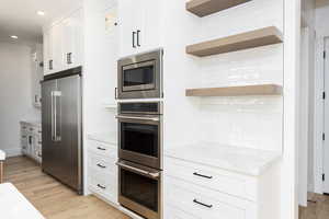 Kitchen with stainless steel appliances, white cabinets, light wood-type flooring, light stone countertops, and open shelves