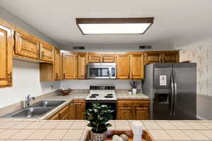 Kitchen featuring wallpapered walls, appliances with stainless steel finishes, brown cabinetry, and tile countertops