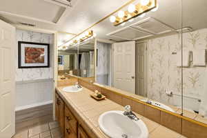 Full bath with wallpapered walls, a textured ceiling, double vanity, and a wainscoted wall