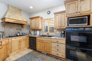 Kitchen featuring black appliances, premium range hood, light countertops, decorative backsplash, and light brown cabinetry