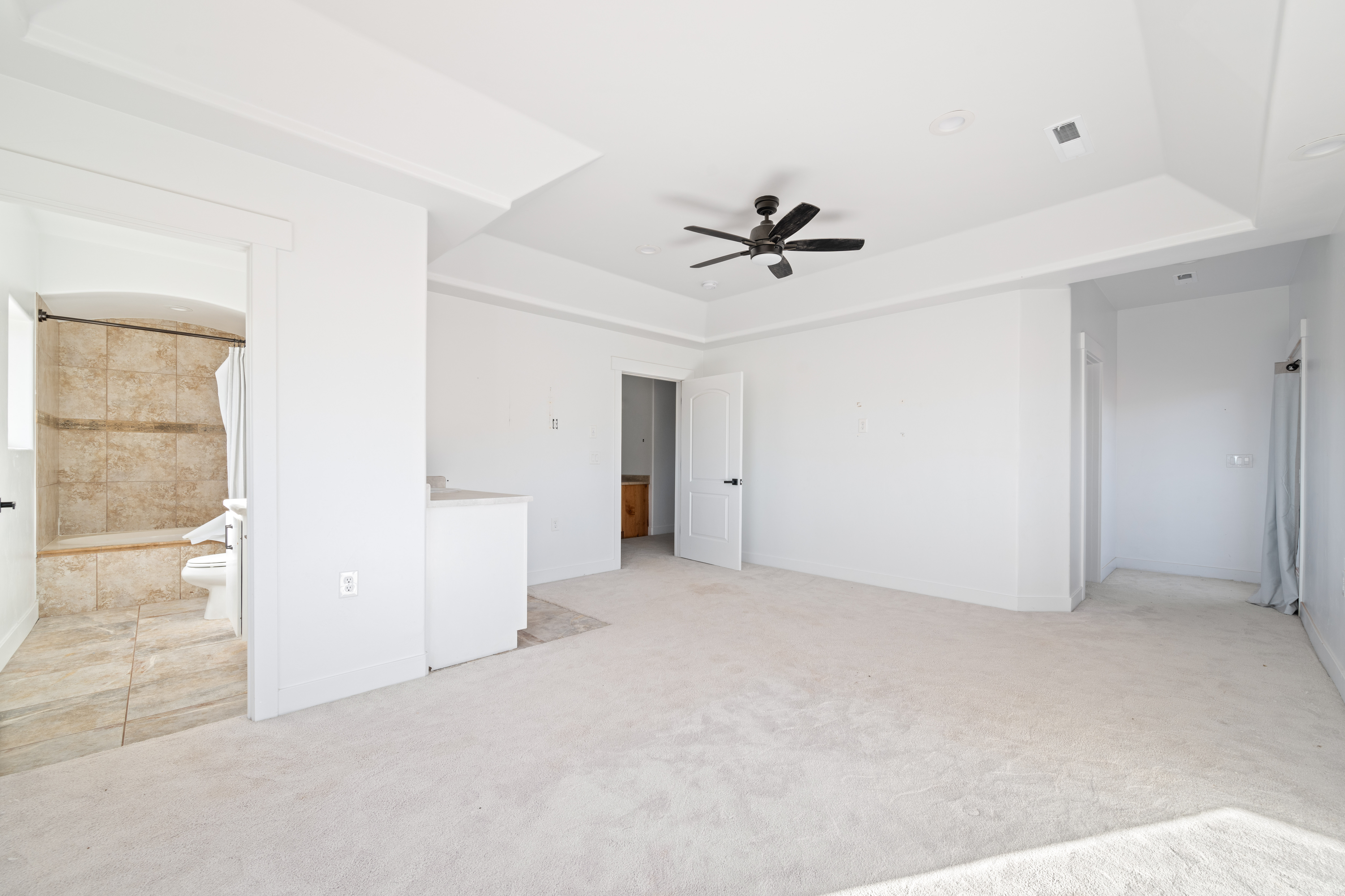 Unfurnished bedroom featuring light colored carpet, ceiling fan, ensuite bath, and a tray ceiling