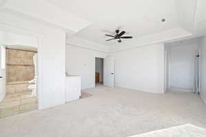 Unfurnished bedroom featuring light colored carpet, ceiling fan, ensuite bath, and a tray ceiling