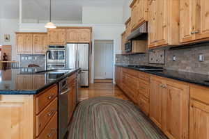 Kitchen with decorative backsplash, dark stone counters, hanging light fixtures, appliances with stainless steel finishes, and light wood finished floors