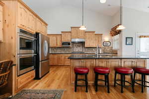 Kitchen featuring hanging light fixtures, stainless steel appliances, an island with sink, high vaulted ceiling, and light brown cabinets