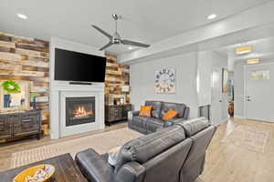 Living room with light wood finished floors, a ceiling fan, wooden walls, a glass covered fireplace, and recessed lighting