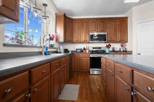 Kitchen with appliances with stainless steel finishes, brown cabinetry, dark wood-type flooring, and decorative light fixtures