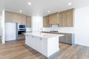 Kitchen with stainless steel appliances, a center island with sink, backsplash, light wood-style floors, and light stone counters