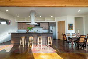 Kitchen featuring dark stone counters, a breakfast bar area, modern cabinets, island range hood, and stainless steel fridge