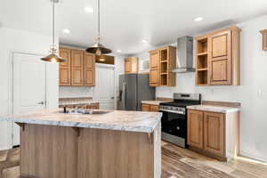 Kitchen with stainless steel appliances, wall chimney range hood, light countertops, a center island with sink, and hanging light fixtures