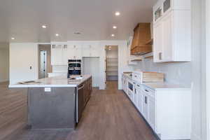 Kitchen featuring glass insert cabinets, white cabinets, an island with sink, recessed lighting, and dark wood-type flooring