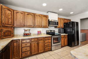 Kitchen featuring appliances with stainless steel finishes, brown cabinetry, light countertops, light tile patterned floors, and a wainscoted wall