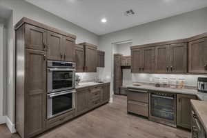 Kitchen featuring double oven, wine cooler, light wood-style floors, dark brown cabinetry, and light stone counters