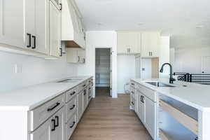 Kitchen featuring light stone counters, light wood-style flooring, a kitchen island with sink, custom exhaust hood, and white cabinetry