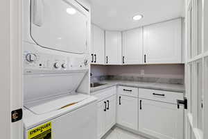 Washroom featuring cabinet space, stacked washer / dryer, light marble finish floors, and recessed lighting