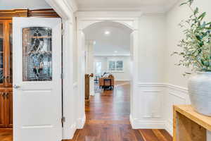 Corridor with arched walkways, dark wood finished floors, crown molding, wainscoting, and a decorative wall
