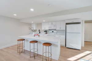 Kitchen with white appliances, white cabinets, light wood-type flooring, a breakfast bar area, and recessed lighting