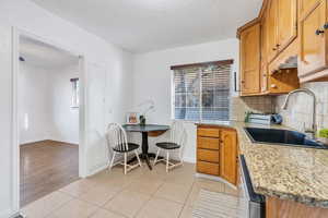 Kitchen featuring brown cabinets, light stone countertops, light tile patterned floors, decorative backsplash, and dishwasher