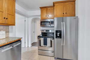 Kitchen featuring appliances with stainless steel finishes, light stone countertops, arched walkways, light tile patterned flooring, and brown cabinetry
