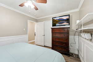 Bedroom with a decorative wall, wainscoting, crown molding, a ceiling fan, and light wood-style flooring