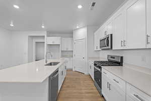 Kitchen with stainless steel appliances, white cabinets, light wood-type flooring, an island with sink, and light stone counters