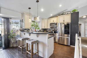 Kitchen featuring cream cabinetry, appliances with stainless steel finishes, dark countertops, vaulted ceiling, and hanging light fixtures