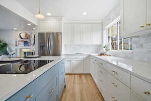 Kitchen featuring stainless steel refrigerator with ice dispenser, light stone countertops, black electric stovetop, blue cabinets, and white cabinetry