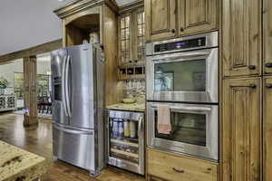 Kitchen with appliances with stainless steel finishes, beverage cooler, glass insert cabinets, brown cabinets, and wood finished floors