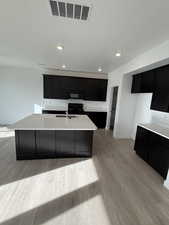 Kitchen with dark cabinetry, an island with sink, black appliances, light wood-style flooring, and recessed lighting